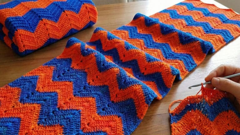 close-up de um trabalho de crochê em andamento sobre uma mesa de madeira clara. A peça utiliza a técnica "Ponto Chama", apresentando um padrão de ziguezague ondulado e fluido. As cores se alternam em listras horizontais de azul escuro, e laranja. No canto inferior direito, uma mão segura uma agulha de crochê metálica, finalizando uma das fileiras.