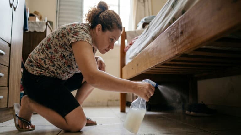 Mulher borrifando mistura com vinagre e água embaixo da cama após a limpeza de casa