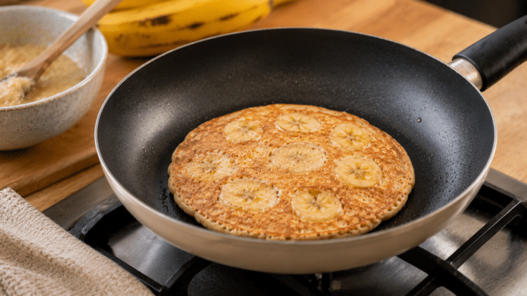 Panqueca de banana sendo preparada na frigideira, com rodelas da fruta incorporadas à massa de forma natural e dourada.