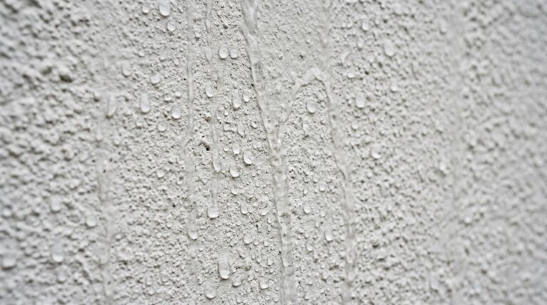 Close-up de uma parede branca com textura rústica, evidenciando o efeito hidrofóbico onde dezenas de gotas de água repuxam e escorrem sem serem absorvidas pela superfície.