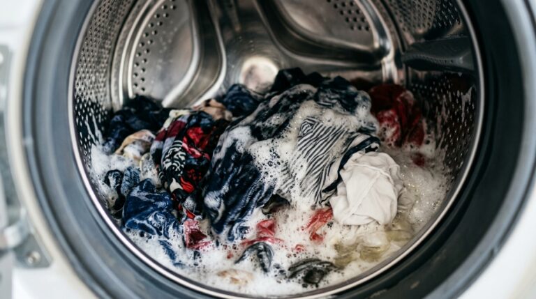 Visão interna do tambor de aço inox de uma máquina de lavar frontal em funcionamento. Roupas coloridas, incluindo peças azuis e listradas, estão envoltas em água e espuma branca durante o ciclo de lavagem.
