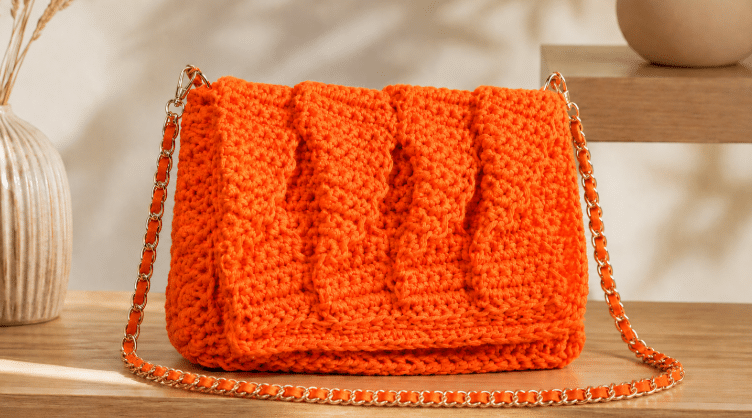Bolsa de crochê laranja vibrante com textura em relevo e alça de corrente completa, exposta sobre uma superfície de madeira em ambiente minimalista e elegante.