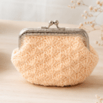 Bolsinha moedeiro de crochê em tom pêssego claro, com textura artesanal em ponto fantasia e fecho metálico prateado, apoiada sobre mesa clara com detalhes delicados ao redor.