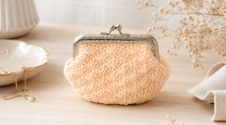 Bolsinha moedeiro de crochê em tom pêssego claro, com textura artesanal em ponto fantasia e fecho metálico prateado, apoiada sobre mesa clara com detalhes delicados ao redor.