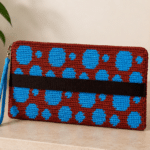 Bolsa carteira de crochê marrom com bolinhas azuis, alça lateral azul e faixa preta horizontal sobre superfície clara em ambiente minimalista.