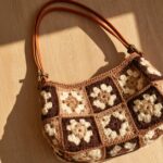 Bolsa feita com quadradinhos de crochê em tons de marrom e bege, com motivos florais e alças compridas.