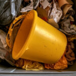 Balde amarelo descartado dentro de um recipiente com panos, sacolas e outros materiais usados.