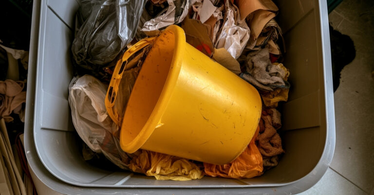 Balde amarelo descartado dentro de um recipiente com panos, sacolas e outros materiais usados.