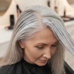 Mulher de cabelos grisalhos recebe aplicação de tonalizante nos fios em um salão de beleza.
