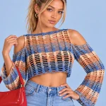Mulher usa blusa cropped de crochê vazada em azul e bege, com mangas longas e ombros abertos, combinada com jeans.
