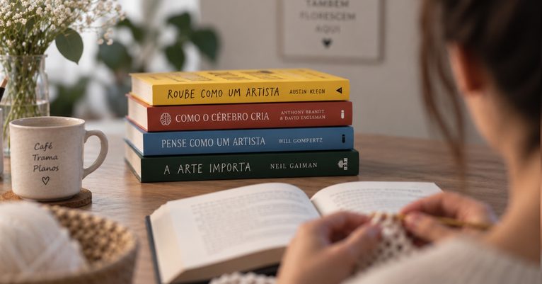 Mulher fazendo crochê enquanto lê, com livros sobre criatividade e arte sobre a mesa.