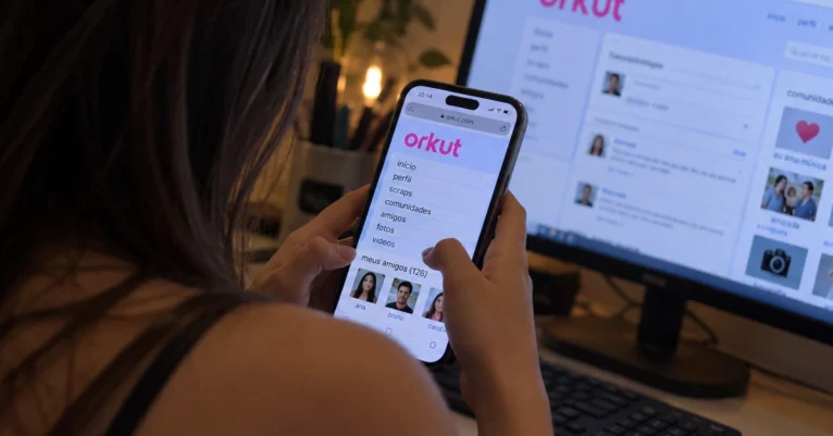 Pessoa acessa uma interface inspirada no Orkut pelo celular, diante de um computador exibindo a rede social.