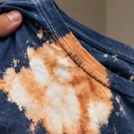 Mão segura camiseta azul com grandes manchas descoloridas provocadas por água sanitária.