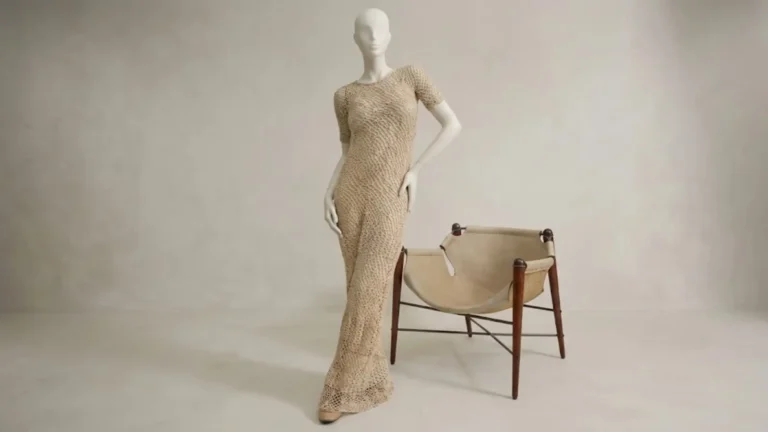 Vestido longo de crochê em tom bege claro com mangas curtas e decote careca, exibido em um manequim branco minimalista ao lado de uma cadeira de couro e madeira em estúdio iluminado.