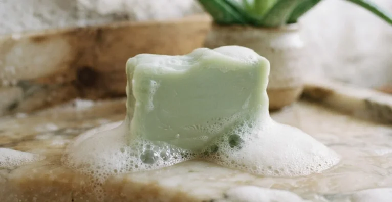 Sabonete artesanal verde de babosa com espuma sobre uma superfície clara e planta de babosa ao fundo.