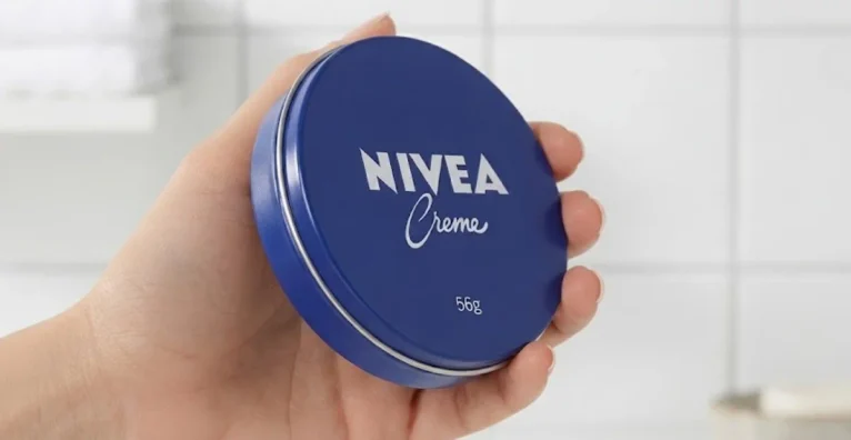 Mão segurando a clássica lata azul do Nivea Creme de 56g em um ambiente de banheiro.